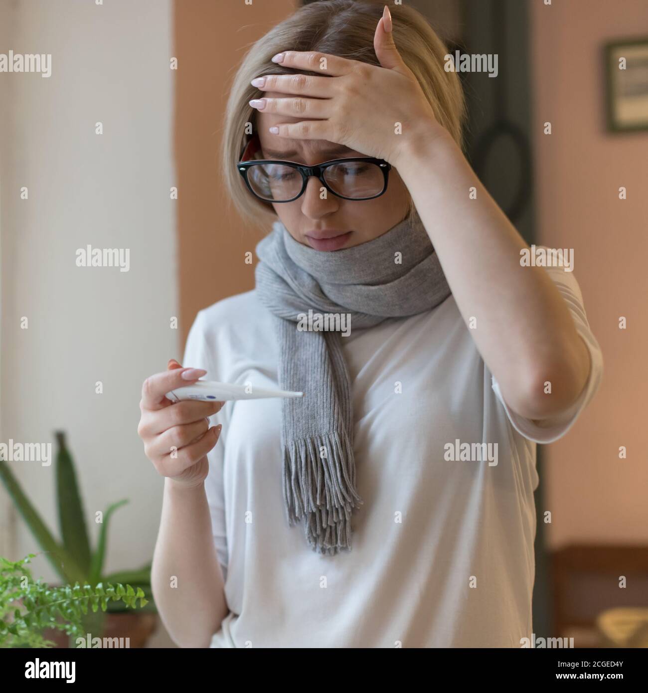 mujer joven en anteojos y camiseta, envuelta en bufanda con síntomas fríos, mirando el termómetro, tocando su frente. Virus, temporada de gripe Foto de stock