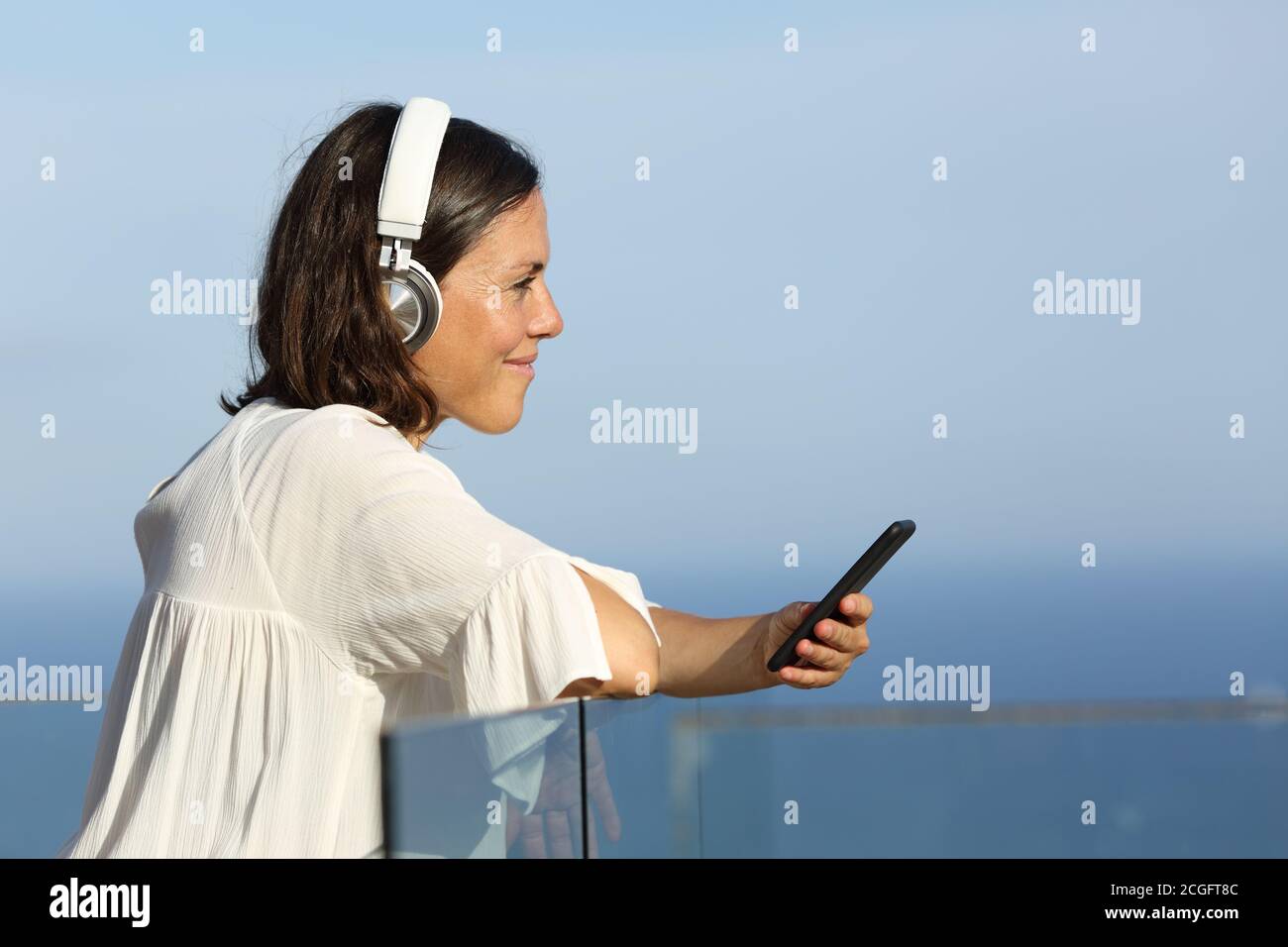 Una mujer adulta con auriculares escucha música en un smartphone de pie en un balcón en la playa Foto de stock