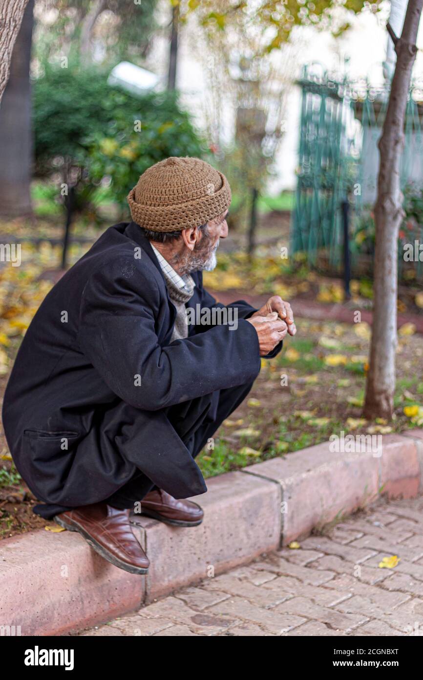 Adana, Turquía, 12/31/2009: Un anciano que lleva sombrero tradicional, abrigo de invierno, y zapatos de cuero se está escurtiendo en el borde de una acera. Está buscando Foto de stock