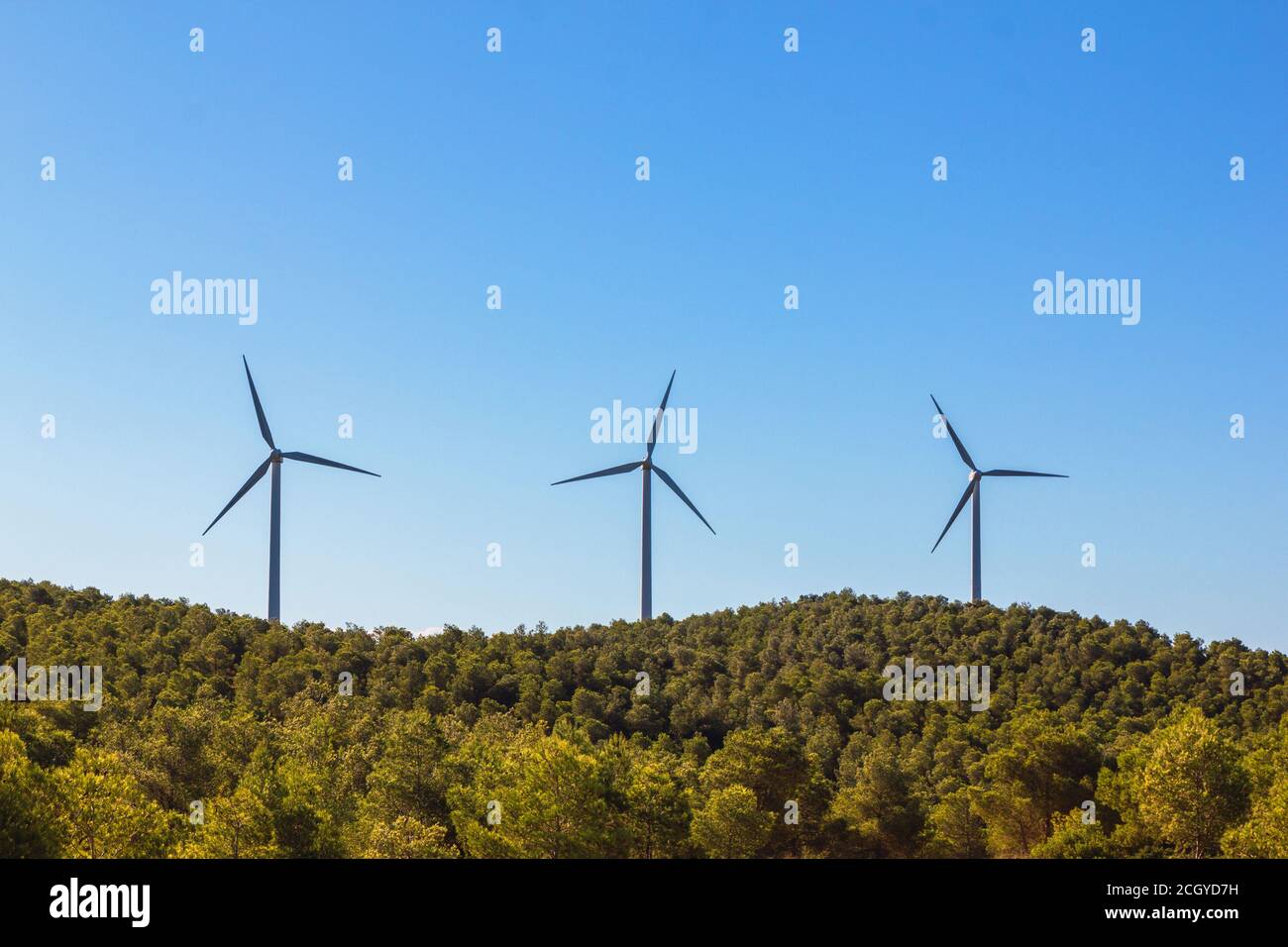 Foto de stock de tres molinos de viento en la cima de una montaña arbolada. Pertenecen a un parque eólico Foto de stock