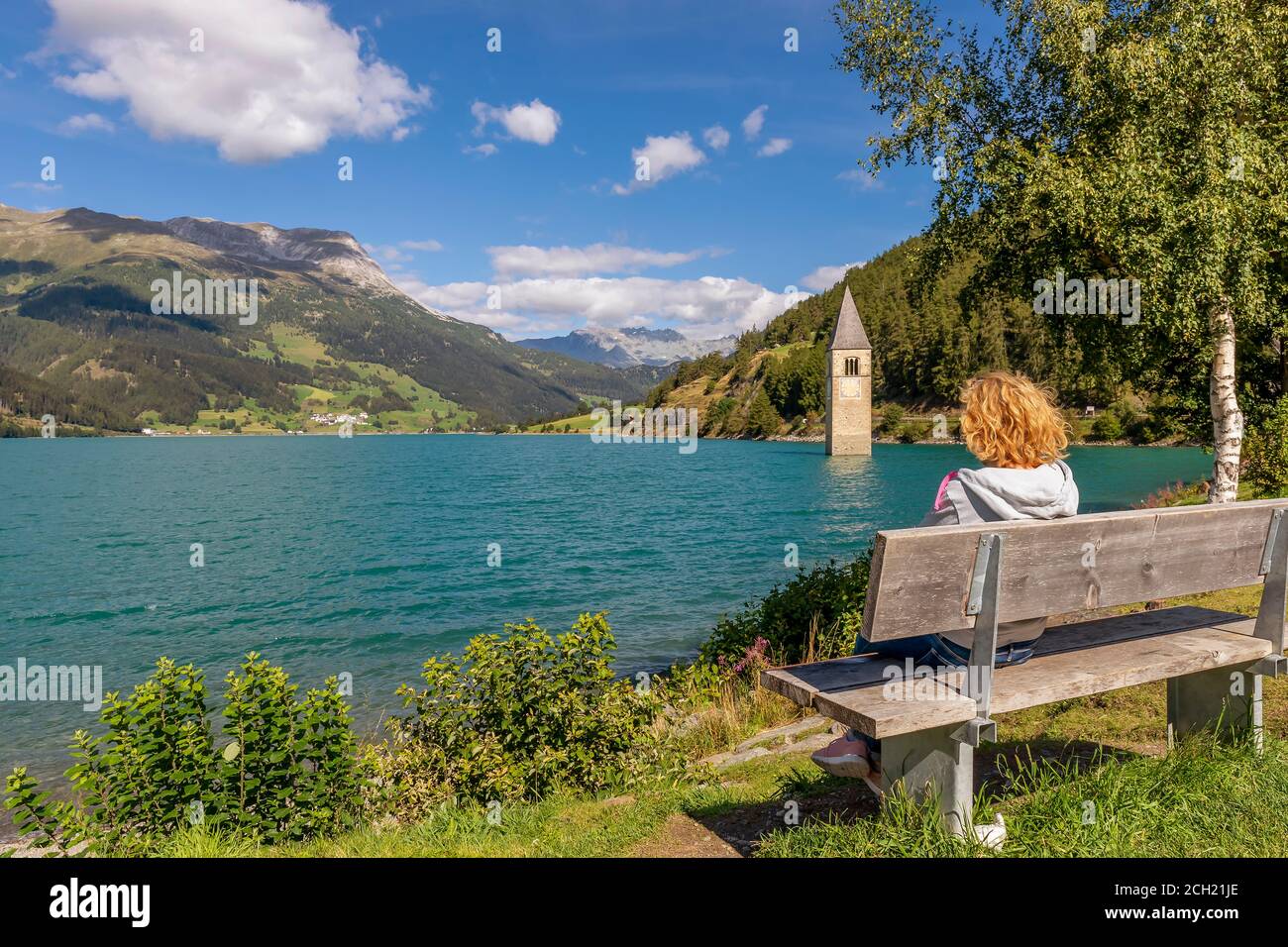 Una rubia sentada en un banco observa el campanario sumergido en el lago Resia, Curon Venosta, Tirol del Sur, Italia Foto de stock