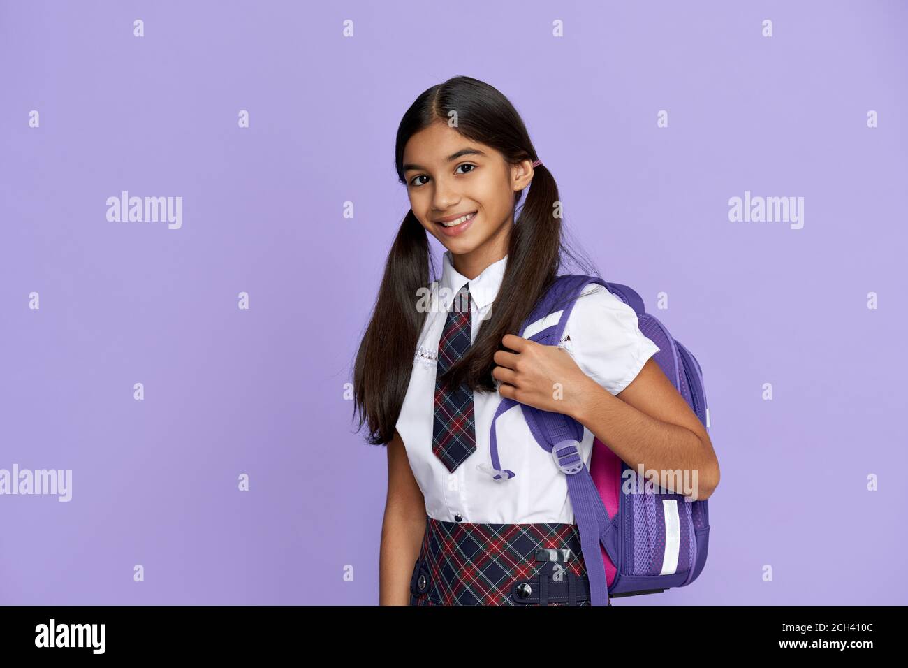 Feliz niña de la escuela india con uniforme sosteniendo la mochila sobre fondo lila. Foto de stock