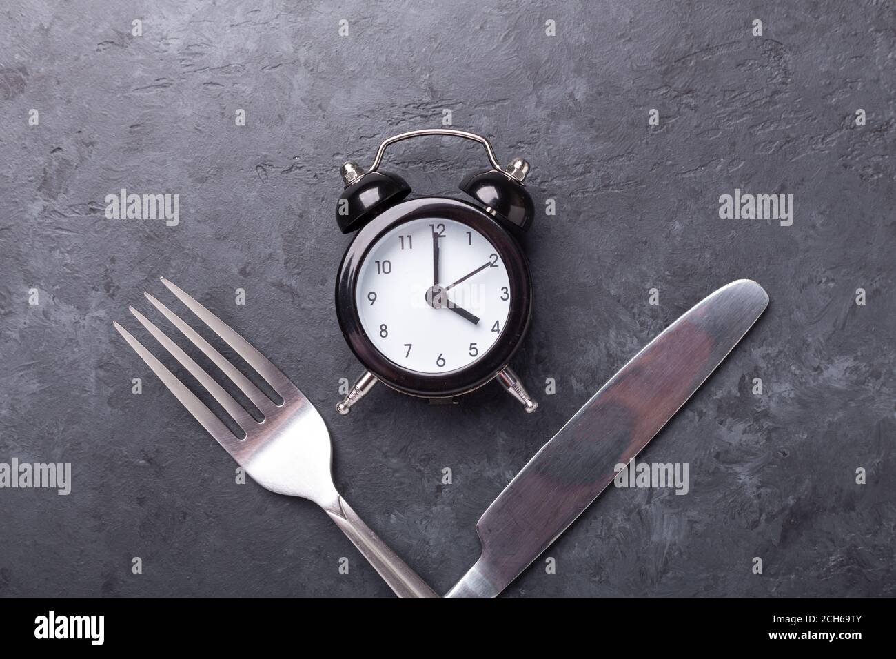 Reloj de alarma negro, tenedor, cuchillo sobre fondo de piedra oscura. Concepto de ayuno intermitente - imagen Foto de stock