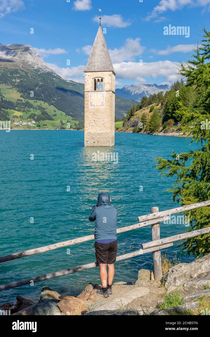 Un hombre vestido deportivo observa el famoso campanario sumergido en el lago Resia en Curon (Graun), Tirol del Sur, Italia Foto de stock