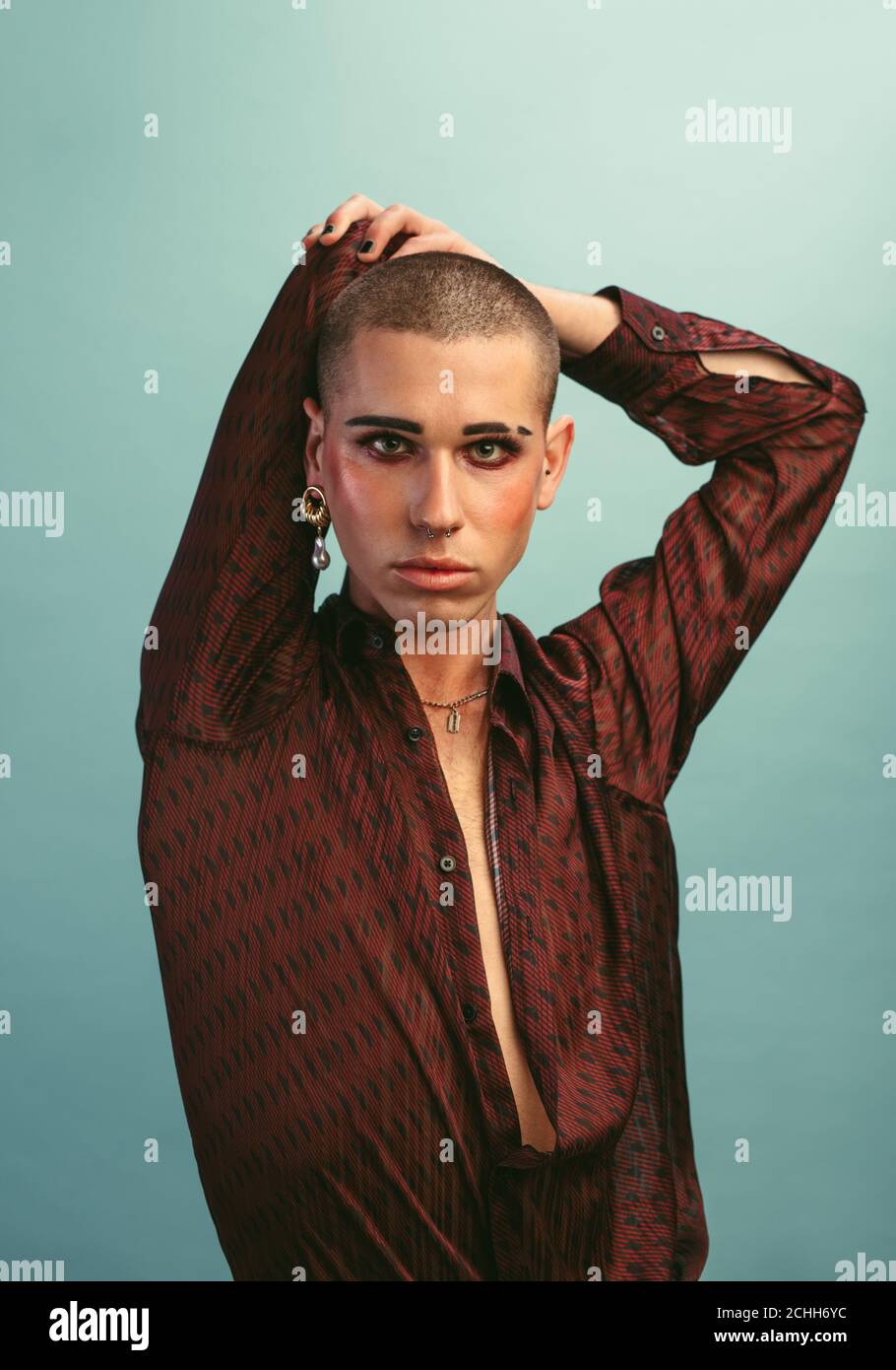 Hombre gay con maquillaje con pendiente de pie en el estudio. Macho transgénero en camisa de seda posando sobre fondo azul. Foto de stock