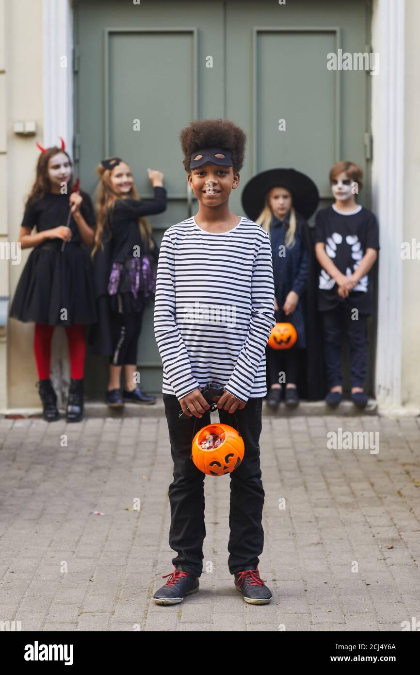 Retrato vertical de toda la longitud de un grupo multiétnico de niños que visten trajes de Halloween mirando la cámara mientras se trampean o tratan juntos, se centran en el niño afroamericano en primer plano Foto de stock