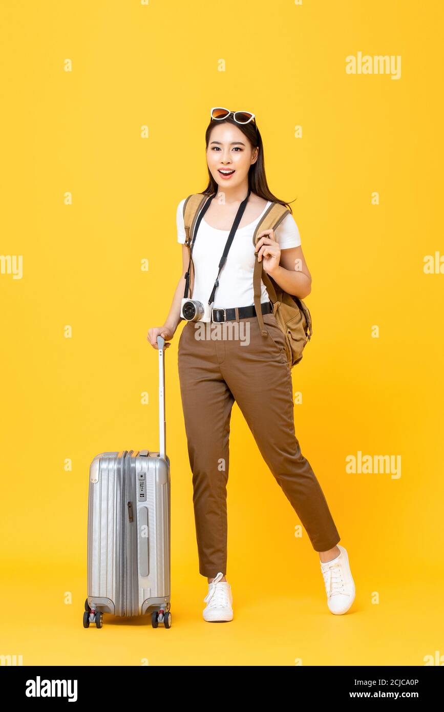 Retrato completo de sonriente mujer joven asiática turista con el equipaje aislado sobre fondo de estudio amarillo Foto de stock