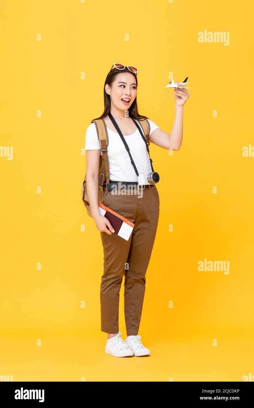 Viaje divertido retrato de joven y sonriente hermosa mujer turística asiática modelo de avión de retención y pasaporte aislados sobre fondo de estudio amarillo Foto de stock