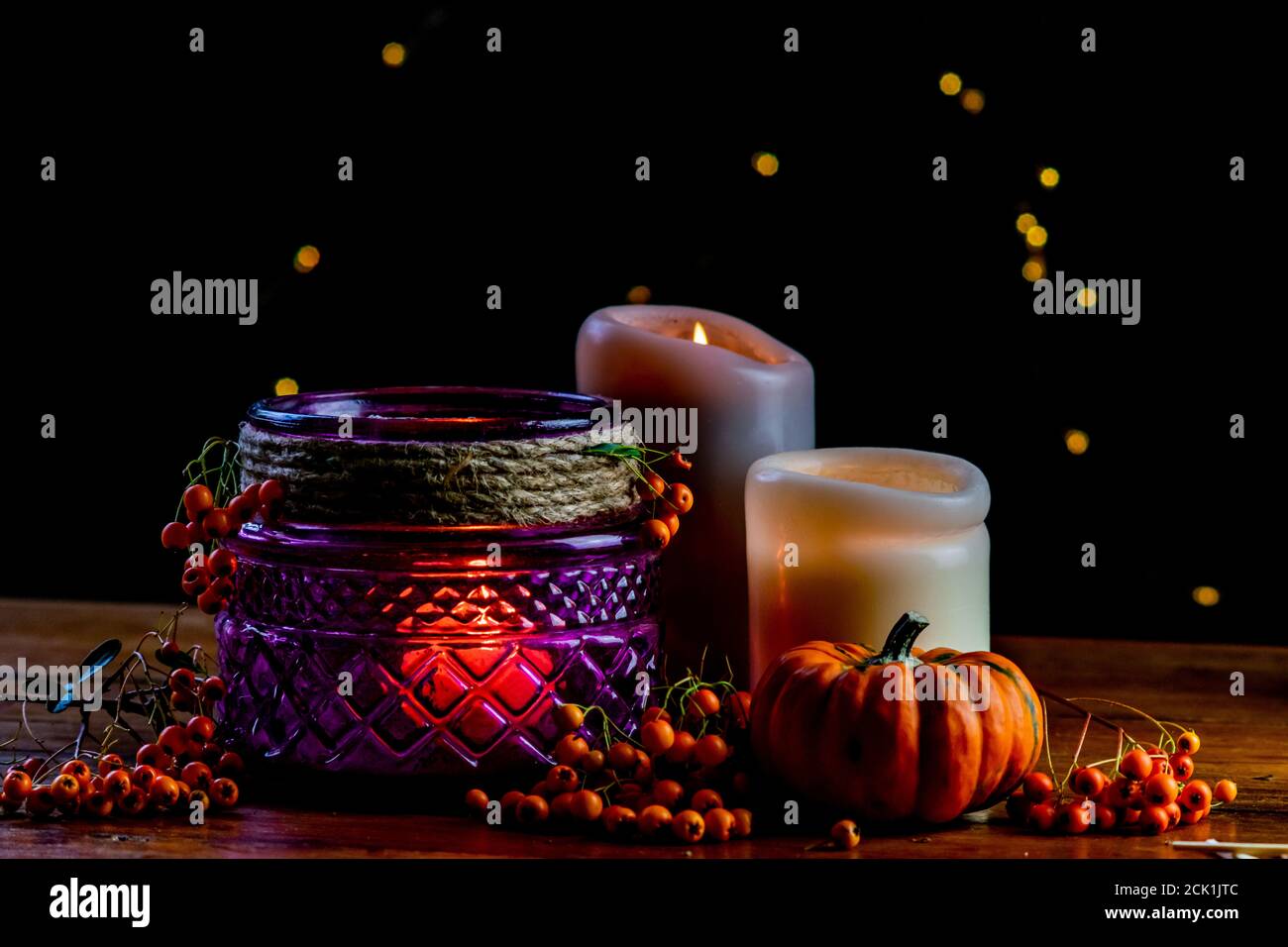 Farol púrpura, naranja dorado pepita calabaza bayas de rowan y dos velas sobre mesa rústica de madera y fondo negro con puntos de bokeh amarillo Foto de stock