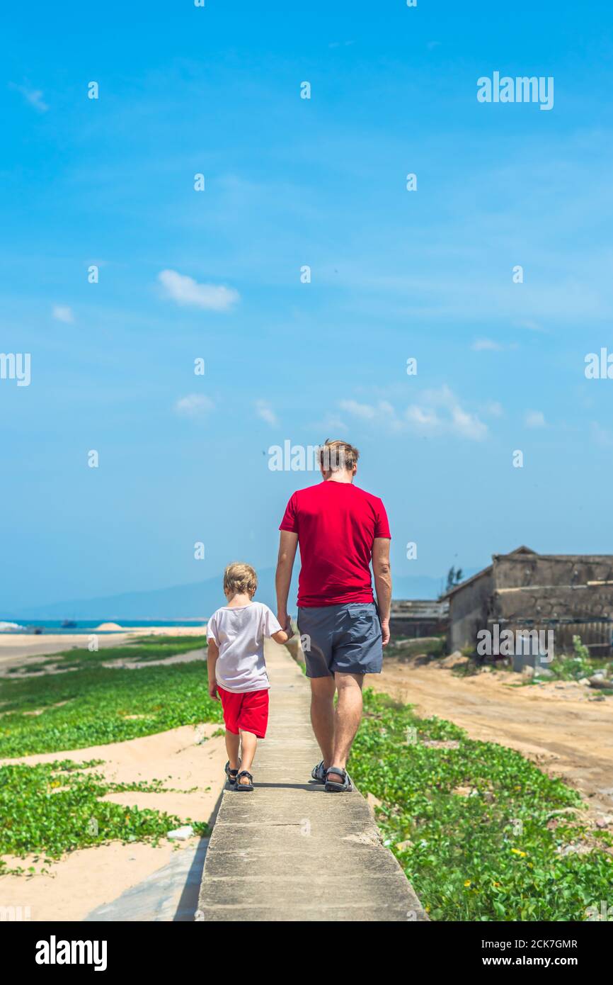 Padre hijo caminar a lo largo de estrecho camino de concreto, mar azul claro cielo verde hierba fondo. Símbolo como papá lleva al niño a la futura edad adulta. Feliz Foto de stock