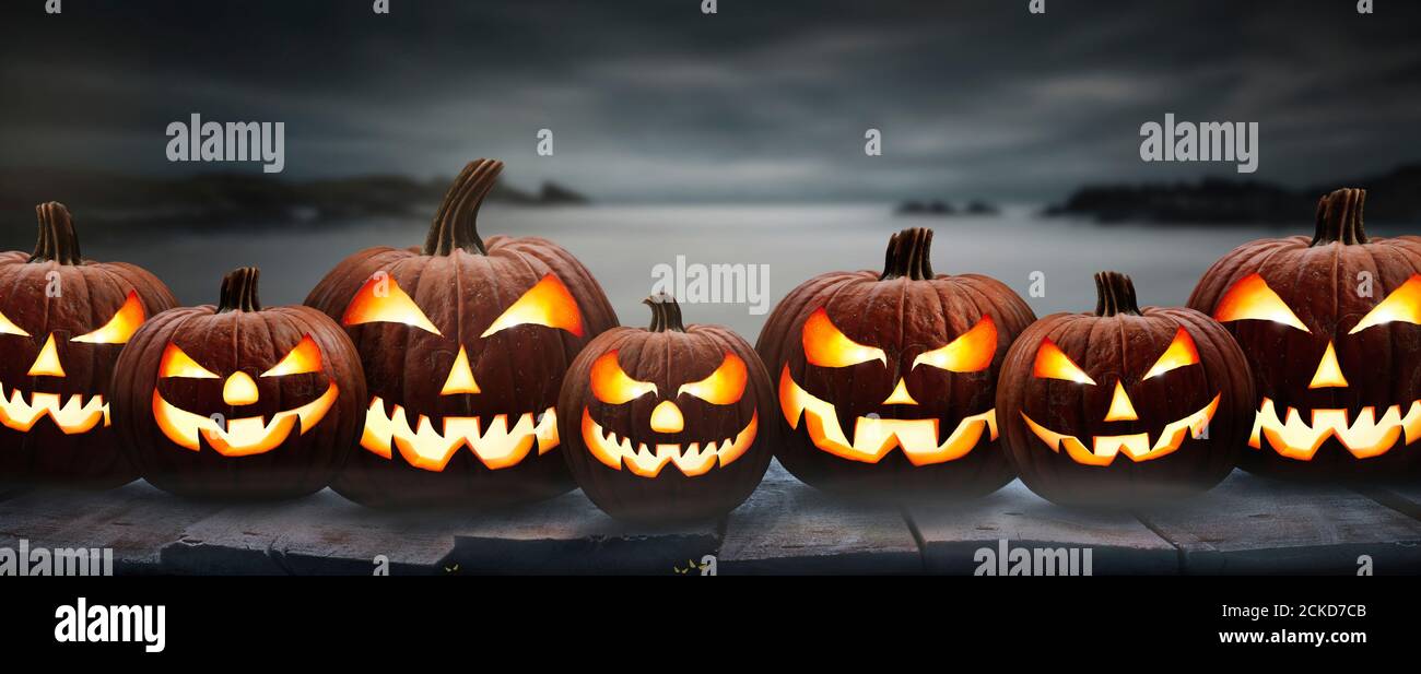 Siete calabazas de halloween, Jack o Lantern, con una cara y ojos malvados en un banco de madera, mesa con un fondo negro gris brumoso de noche costera. Foto de stock