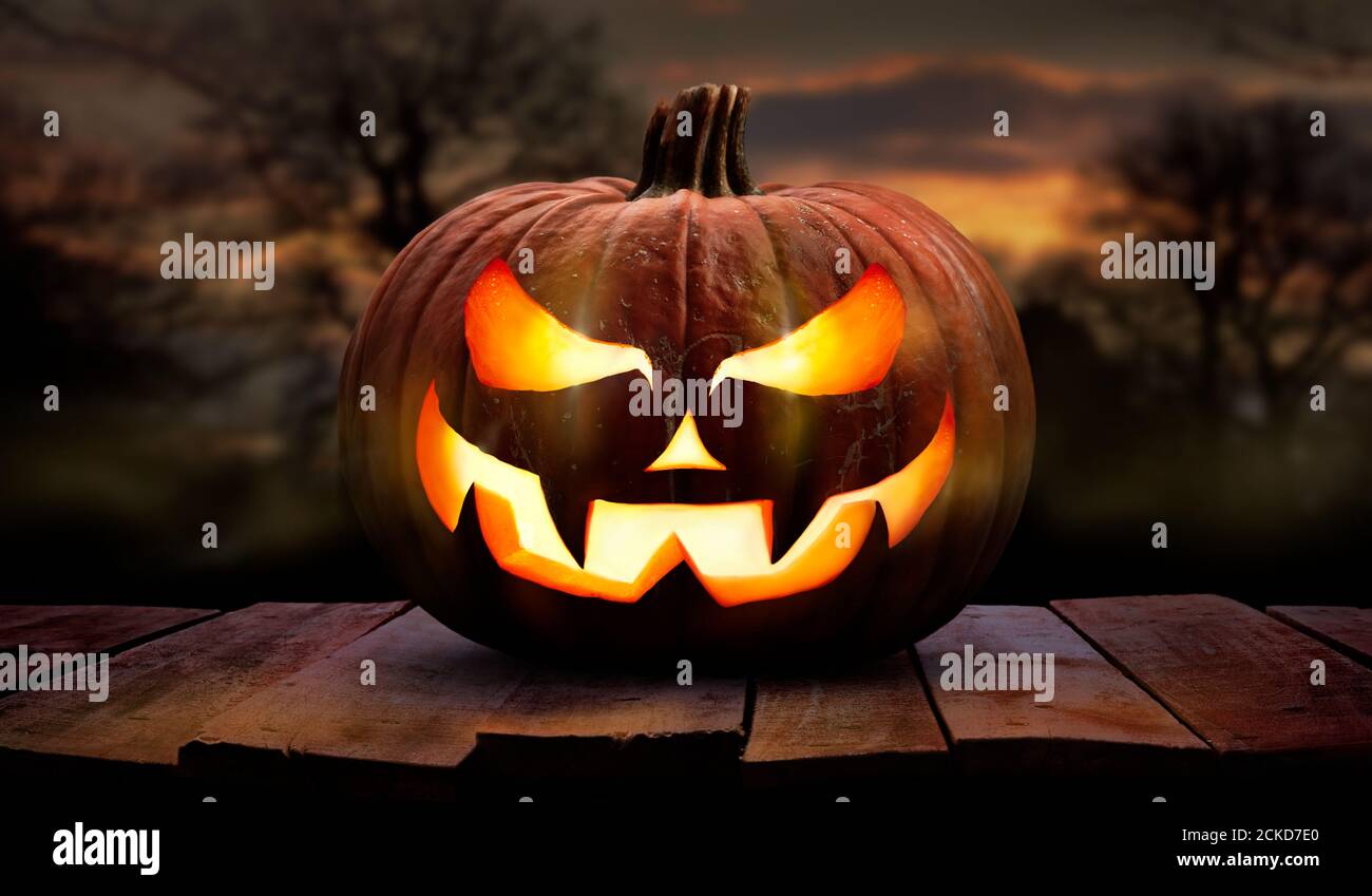 Una calabaza de halloween espeluznante, Jack o Lantern, con un rostro malvado y ojos en un banco de madera, mesa con una puesta de sol, fondo de noche. Foto de stock