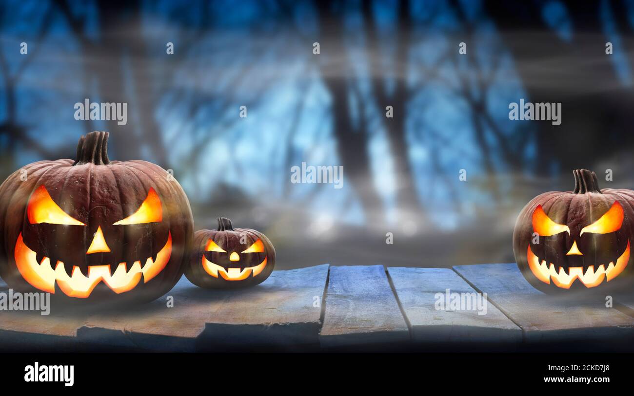 Tres calabazas de halloween espeluznantes, Jack o Lantern, con cara y ojos malvados en un banco de madera, mesa con un fondo de bosque nocturno brumoso con espacio para Foto de stock