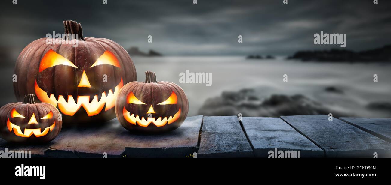 Tres calabazas de halloween, Jack o Lantern, con una cara y ojos malvados en un banco de madera, mesa con un fondo negro gris oscuro de noche con s Foto de stock