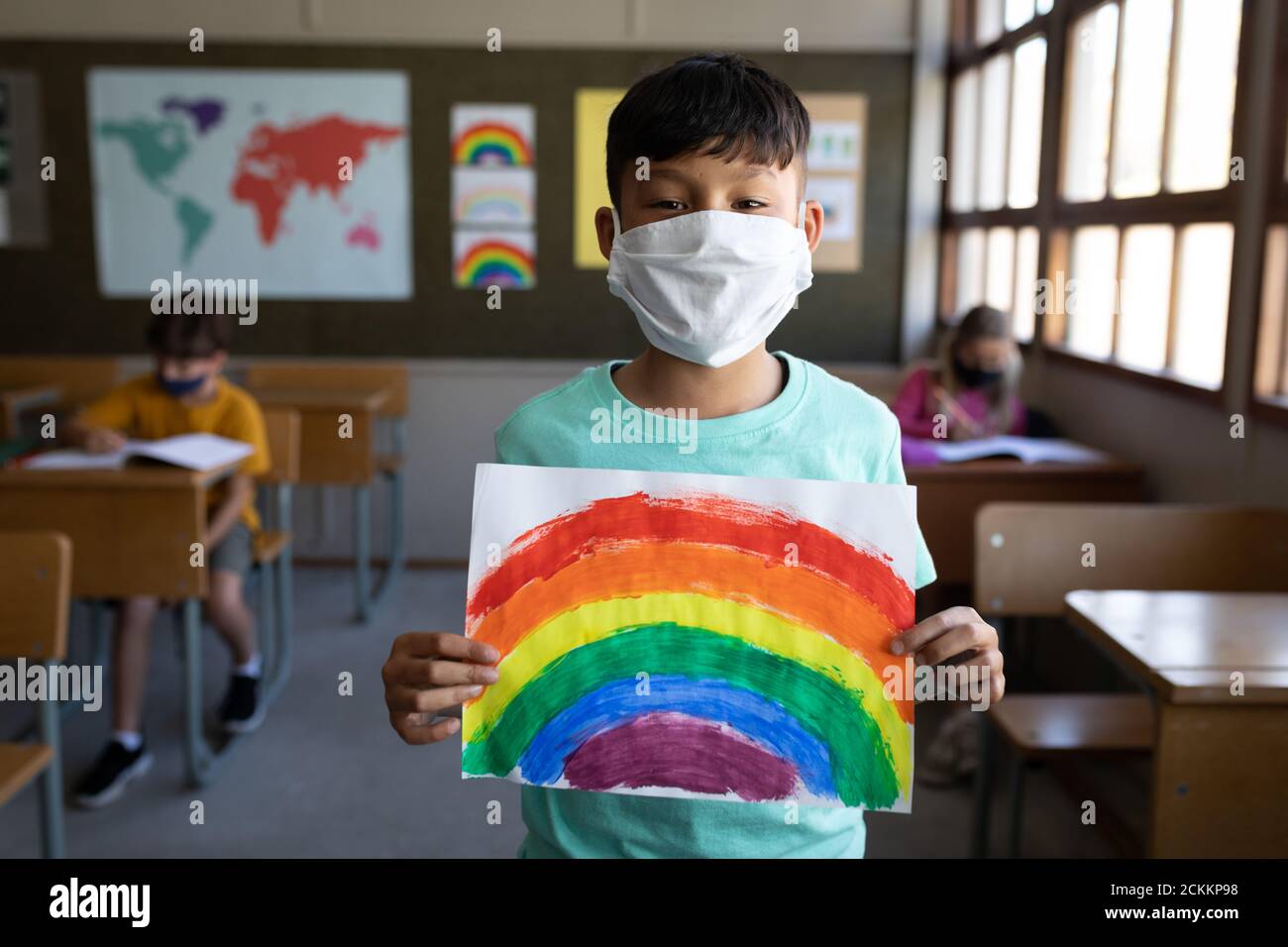 Retrato de niño con máscara de cara sosteniendo un dibujo de arco iris en la escuela Foto de stock