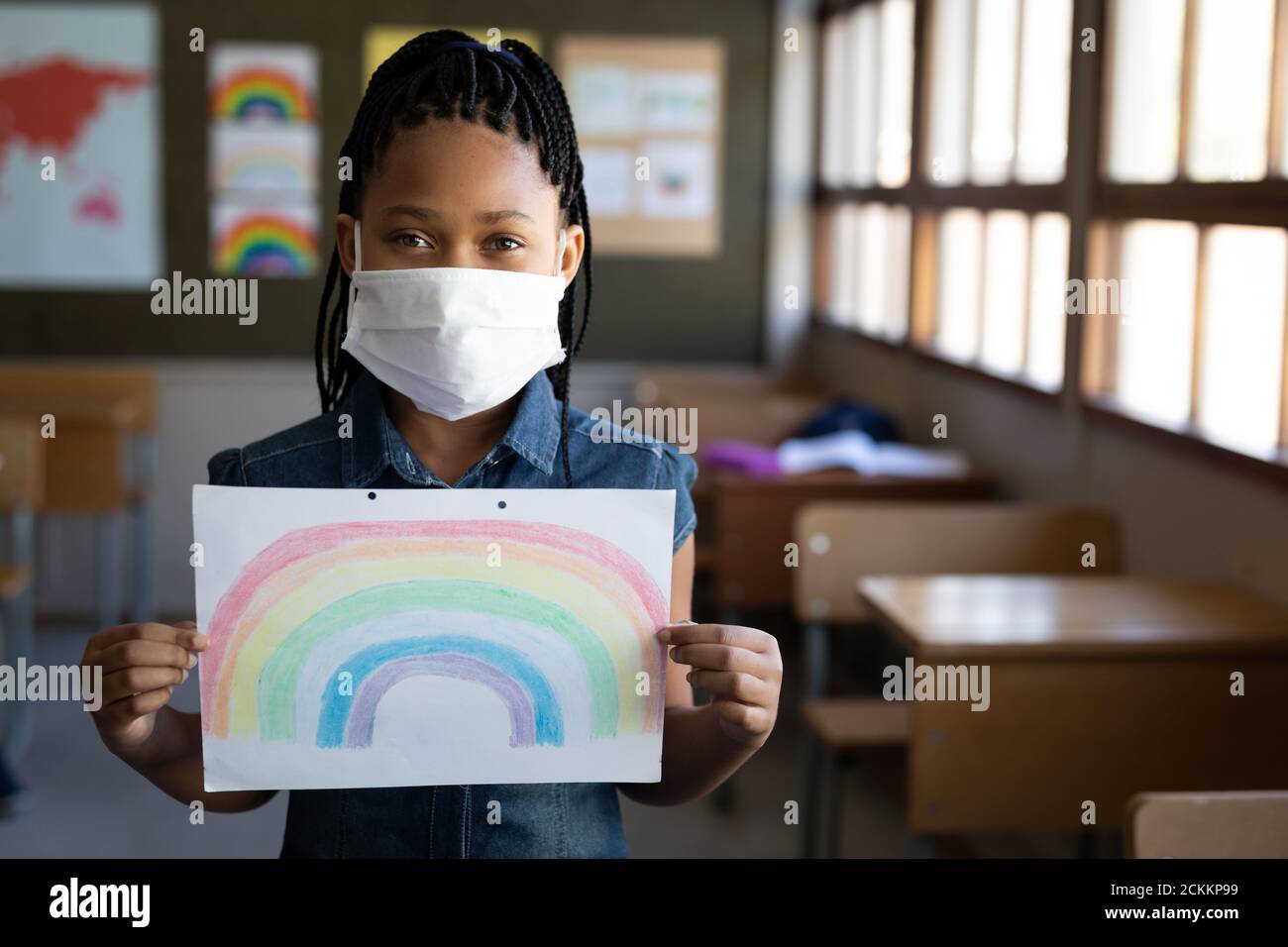 Retrato de una chica con máscara que sostiene un dibujo de arco iris en la escuela Foto de stock