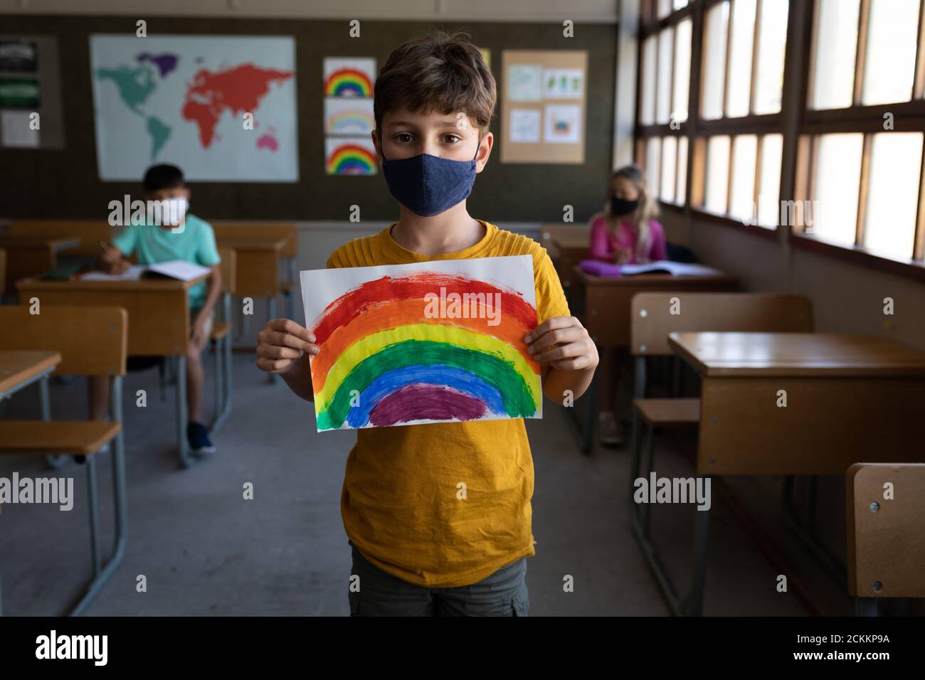 Retrato de niño con máscara de cara sosteniendo un dibujo de arco iris en la escuela Foto de stock