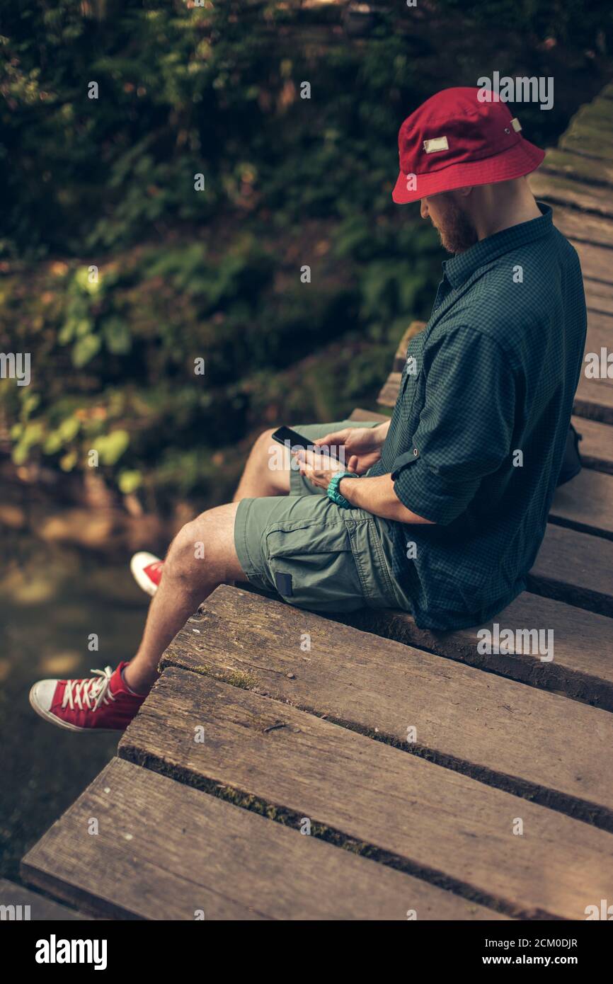 El hombre aventurero perdido en sombrero rojo se sienta en el viejo puente  de madera mientras utiliza el smartphone para descargar el mapa para  encontrar una manera en el bosque. Aventura y