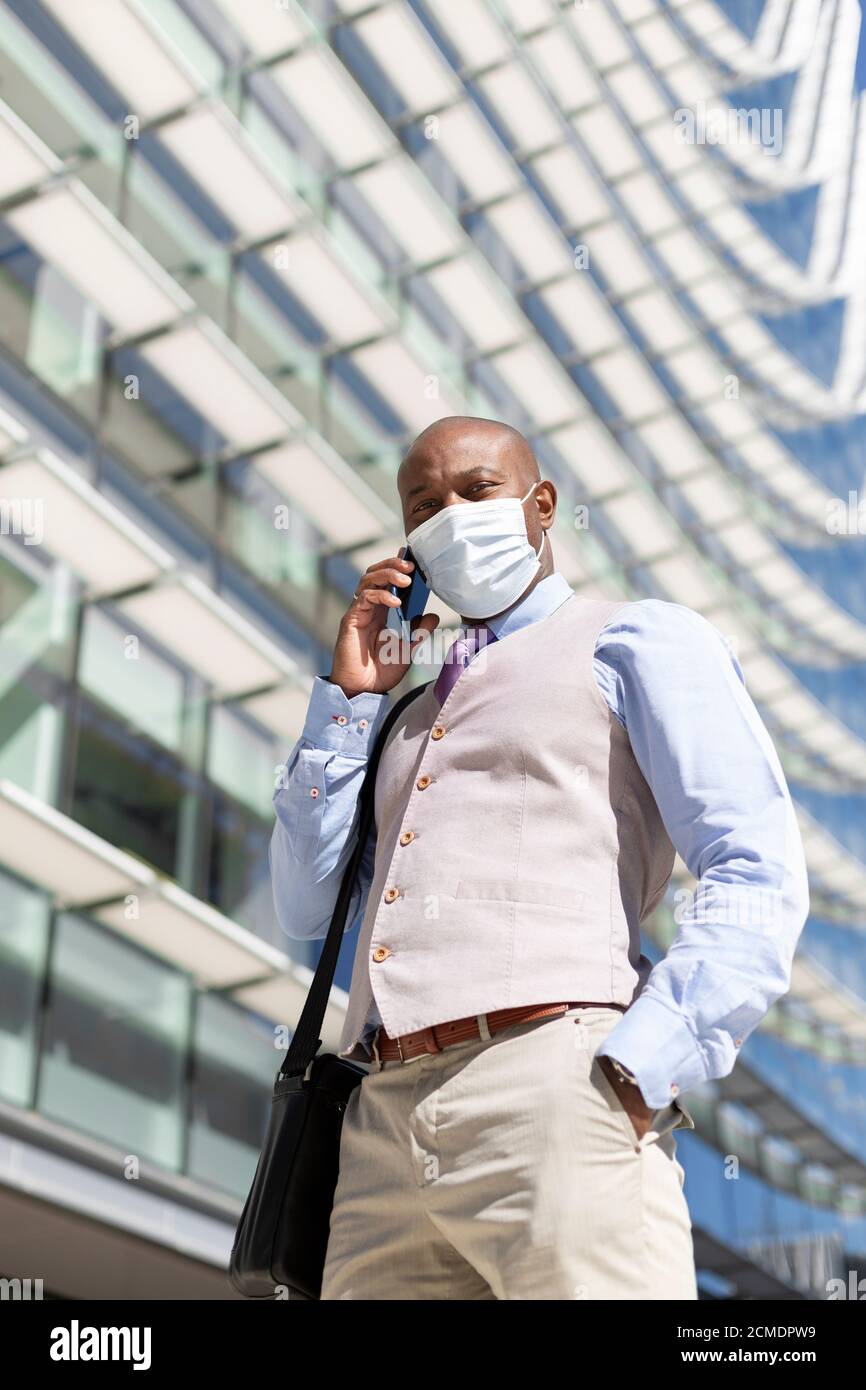 Hombre de negocios negro hablando a través de su smartphone. Está de pie junto a un edificio de oficinas y está usando una máscara. Negocio en el contexto de coronav Foto de stock