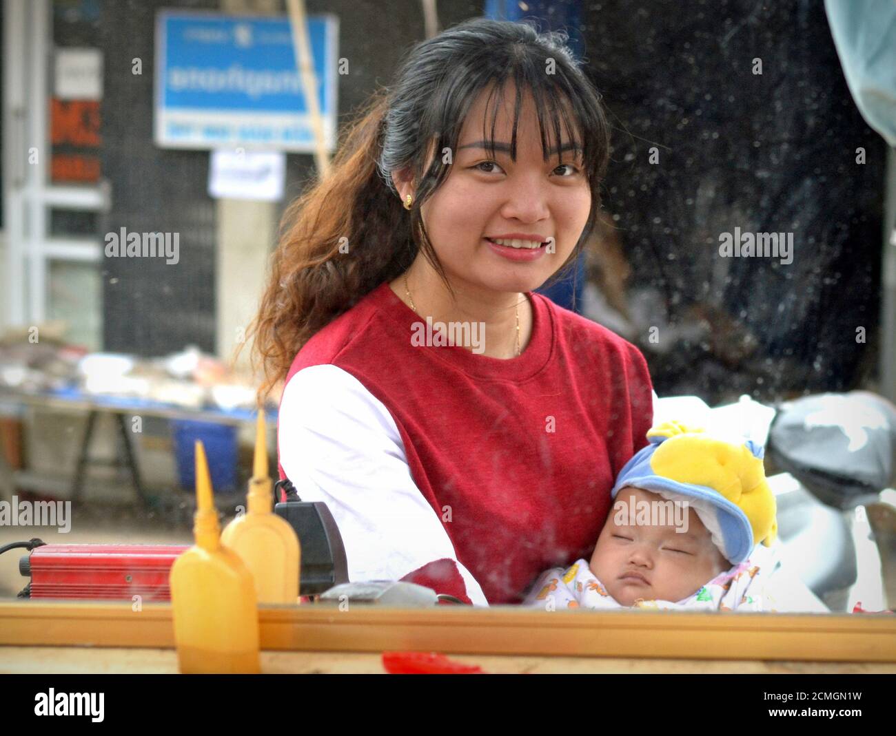 Hermosa joven vietnamita sostiene a su bebé dormido en sus brazos y mira en un sucio y empañado espejo de la peluquería. Foto de stock