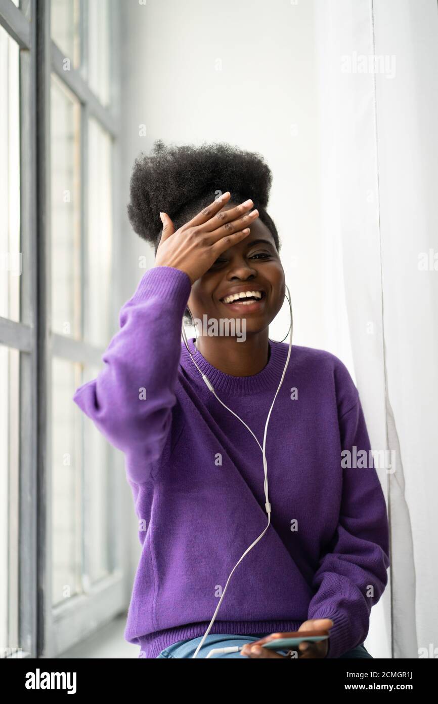 Mujer joven milenaria afroamericana con afro peinado vestir suéter púrpura riendo con una sonrisa de toothy, sentado en el alféizar de la ventana, escucha la música wi Foto de stock