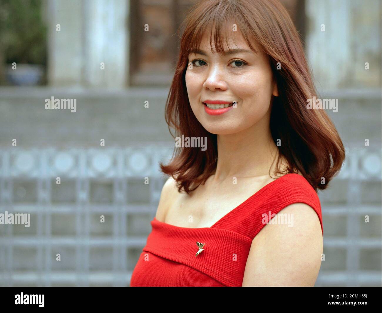 Hermosa joven mujer de negocios vietnamita sonriente con hombro largo pelo marrón lleva un vestido de color rojo a medida y mira la cámara. Foto de stock