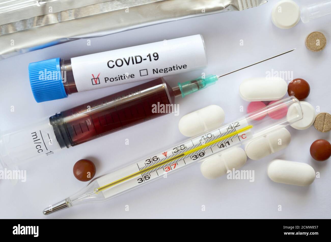 Muestras de sangre en una jeringa desechable y un tubo de ensayo con un resultado positivo para el COVID-19, un termómetro médico de vidrio de mercurio y varias tabletas Foto de stock