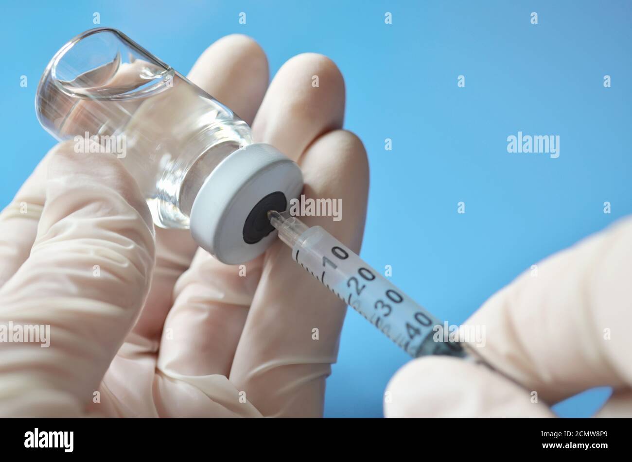 Un profesional médico en guantes de goma extrae una dosis de medicamento de un vial médico utilizando una jeringa desechable sobre un fondo azul claro. Foto de stock