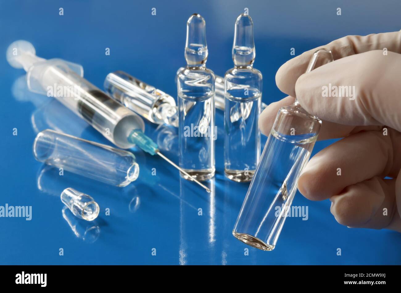 Ampolla con una vacuna en la mano de un médico trabajador en guantes de goma en un fondo azul desenfocado con una jeringa médica y otras ampollas Foto de stock