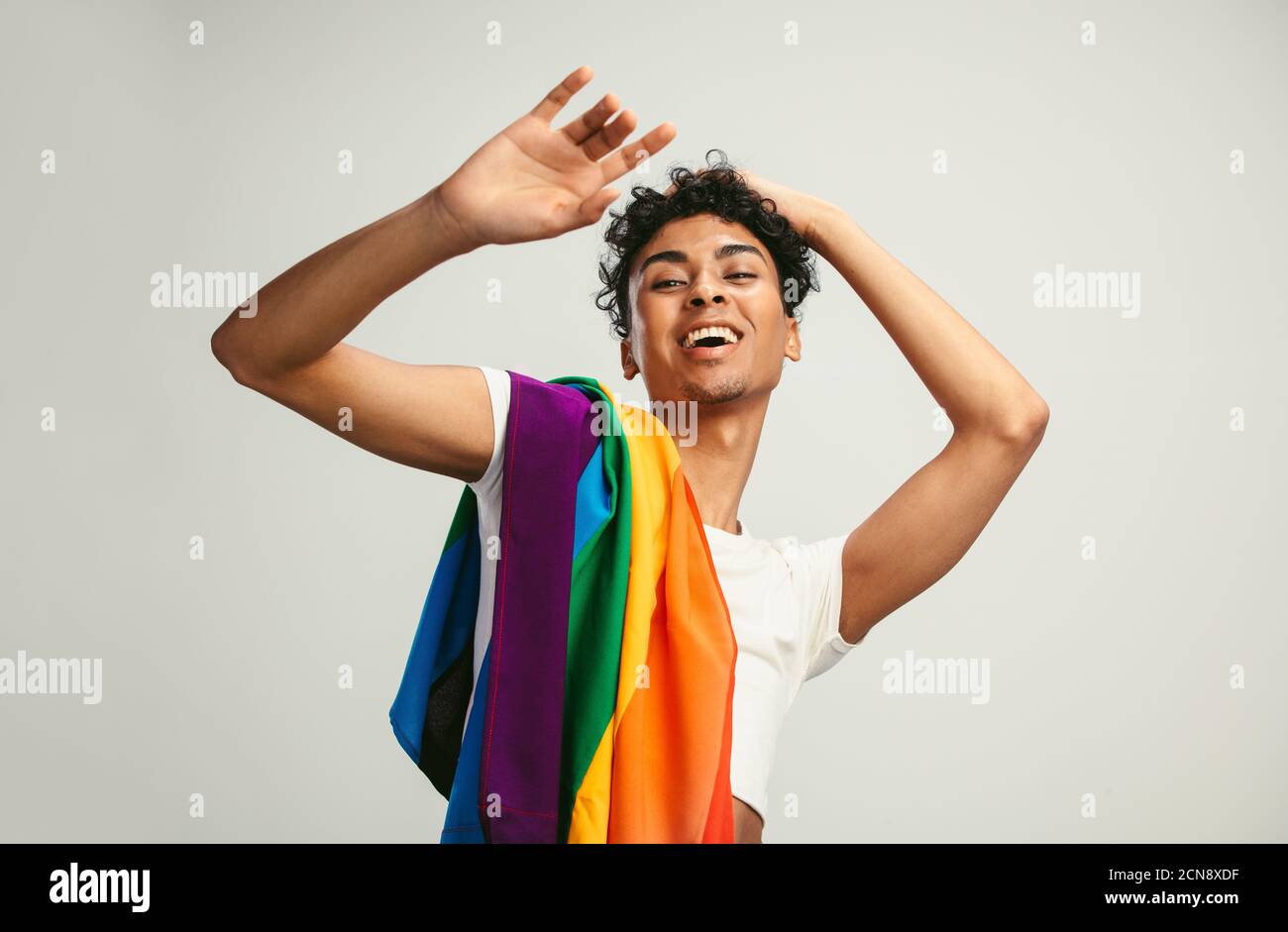 Feliz joven sonriendo con bandera de orgullo sobre fondo blanco. Hombre fluido de género con una bandera lgbtq en su hombro. Foto de stock