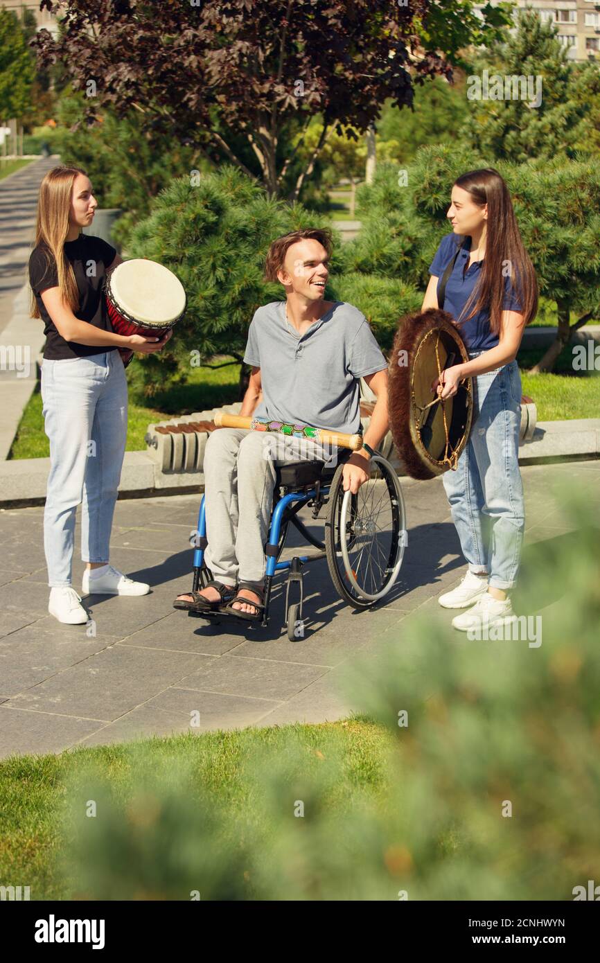 Alegre. Feliz hombre caucásico discapacitado en una silla de ruedas pasar  tiempo con amigos tocando música instrumental en vivo al aire libre.  Concepto de vida social, amistad, posibilidades, inclusión, diversidad  Fotografía de