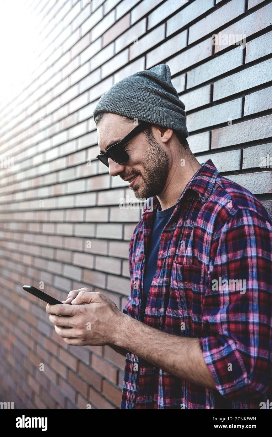 Escribiendo mensaje de texto. Vista lateral de un hombre joven guapo en elegante ropa informal sosteniendo el teléfono móvil mientras se inclina en la pared de ladrillo. Foto de stock
