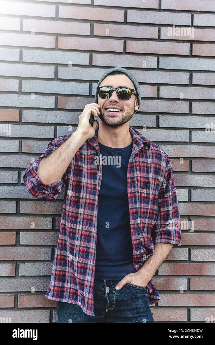 Hablar por teléfono. Vista lateral de un hombre joven guapo con ropa informal y elegante hablando por teléfono móvil en el fondo de la pared de ladrillo. Foto de stock