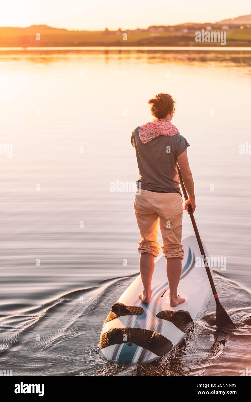 Joven mujer de pie paddleboarding al atardecer, Lago Wallersee, Flachgau, Salzburgo, Austria Foto de stock