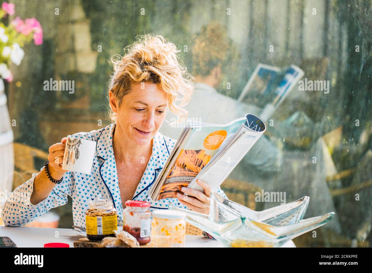Rubia joven madura en pijamas en casa durante el desayuno, leyendo una revista y tomando una taza de café Foto de stock