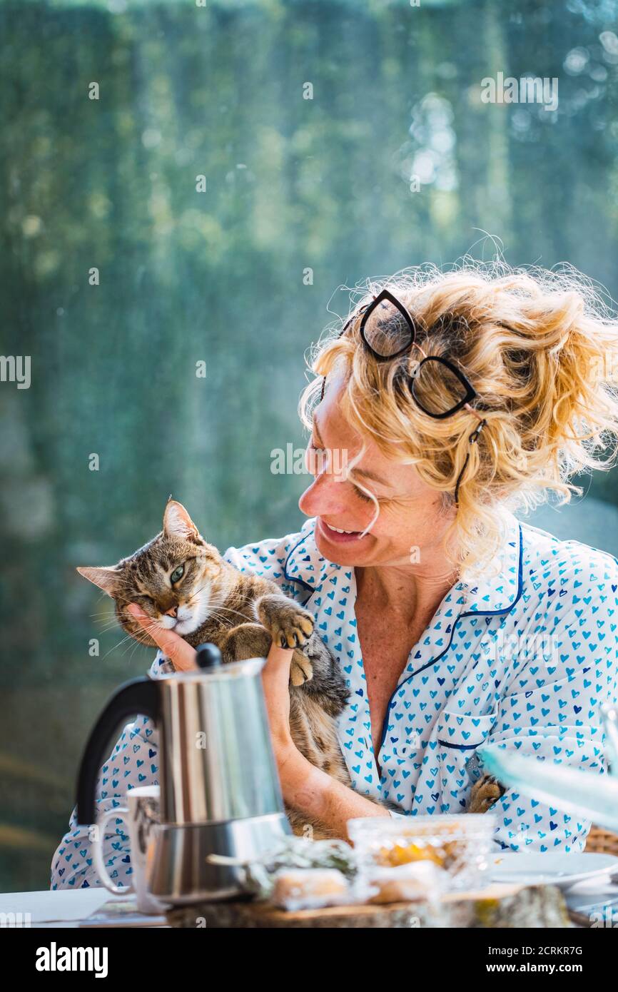Rubia joven madura con gafas en pijama en casa en el desayuno, leyendo una revista y teniendo un momento con su gato. Foto de stock