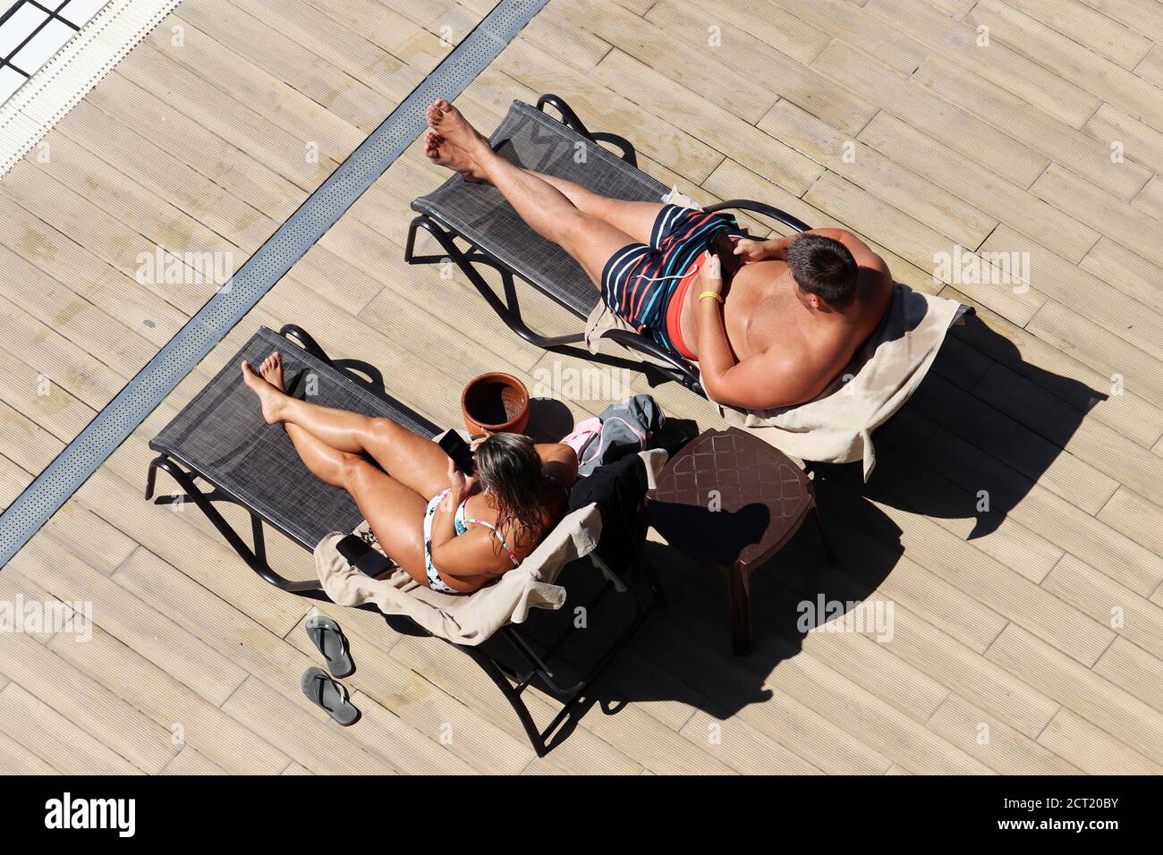 Una pareja tumbada en las tumbonas y tomando el sol cerca de la piscina, vista superior. Mujer en traje de baño y hombre en troncos usando smartphones en una playa Foto de stock