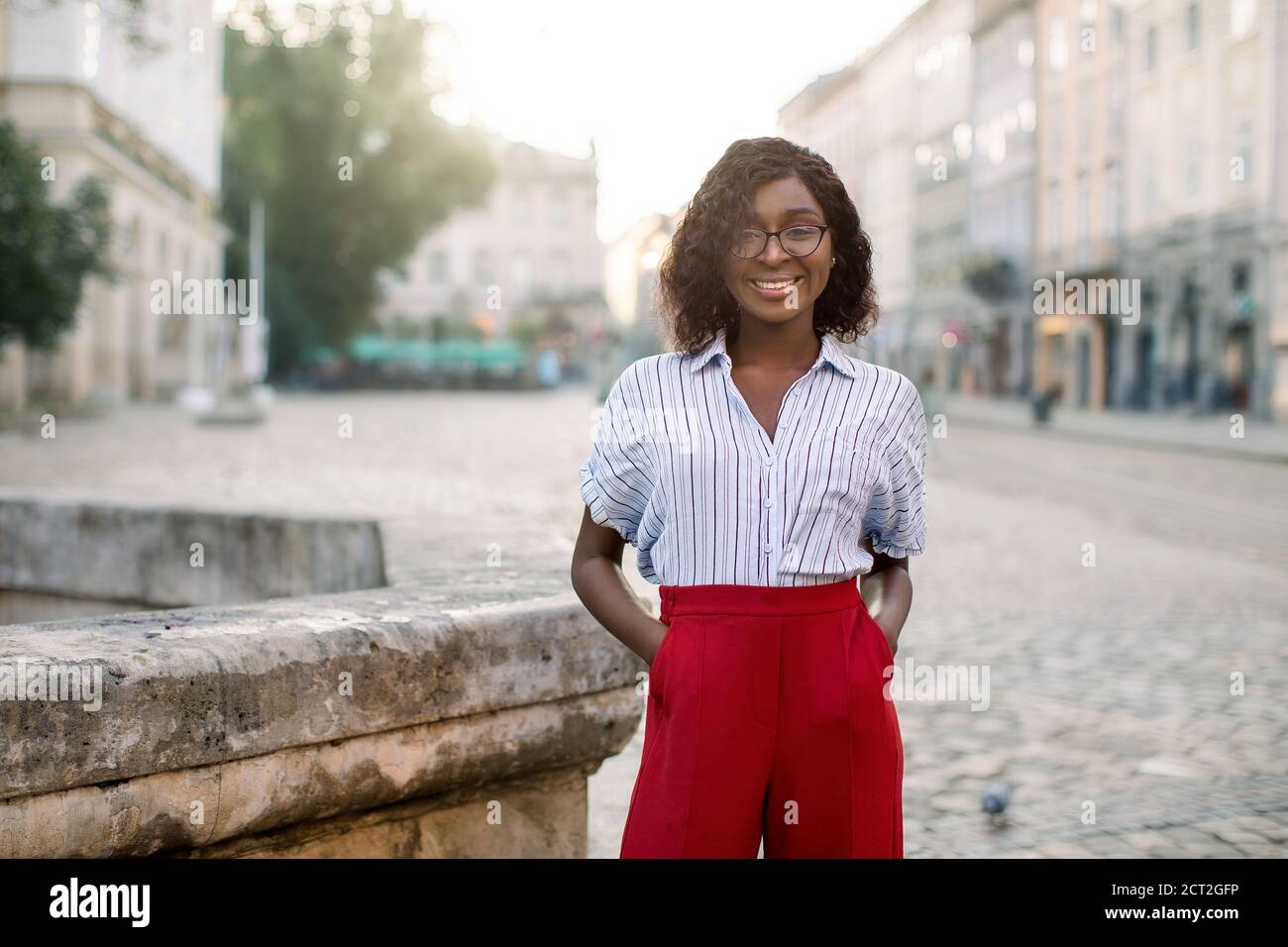 Retrato de sonriente alegre hermosa mujer africana de moda, con gafas,  pantalones rojos y camisa a rayas, de pie en la calle de la ciudad vieja  Fotografía de stock - Alamy