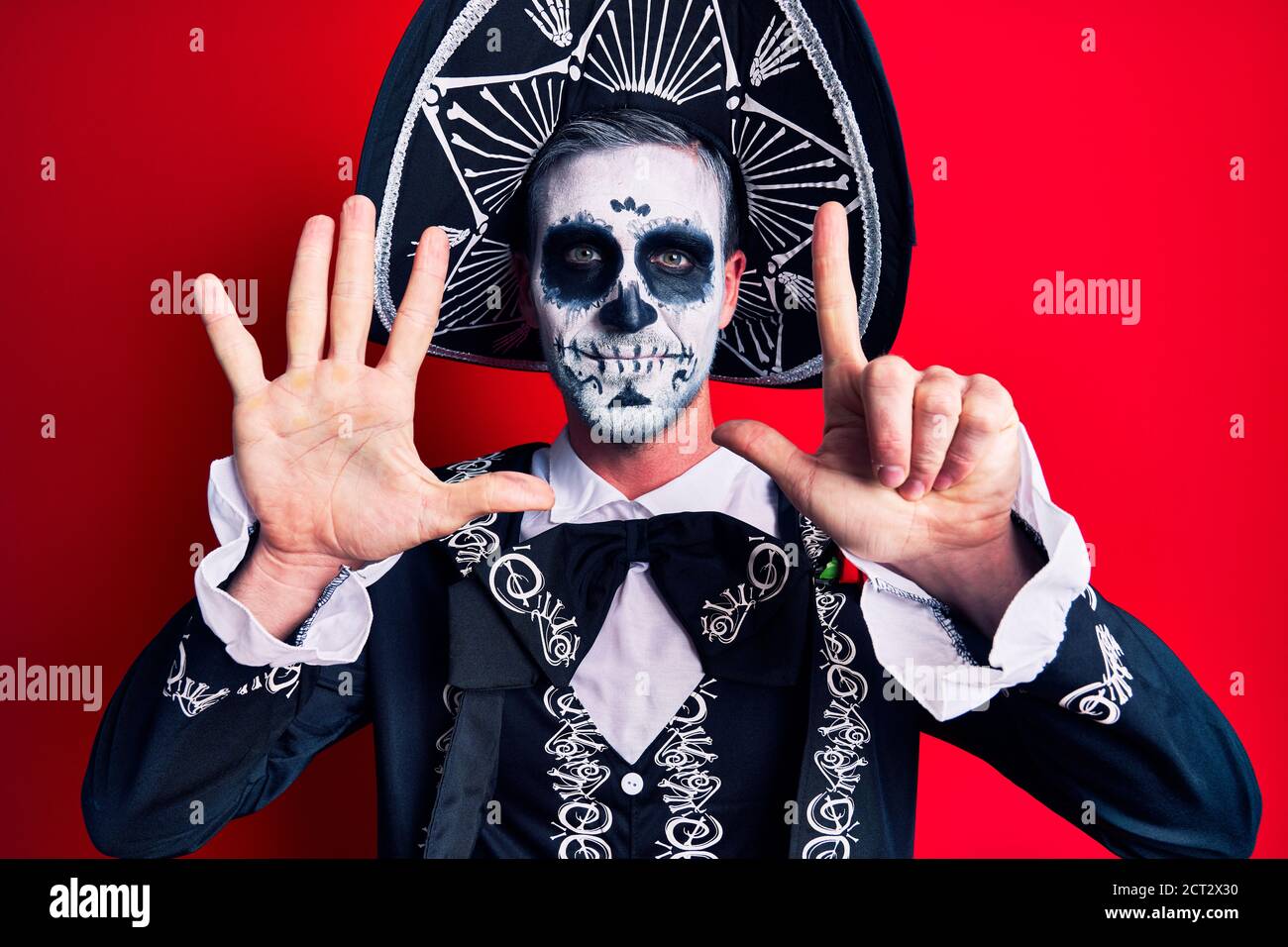 Hombre joven con traje mexicano de día muerto sobre rojo mostrando y señalando con los dedos número siete mientras sonriendo confidente y feliz. Foto de stock