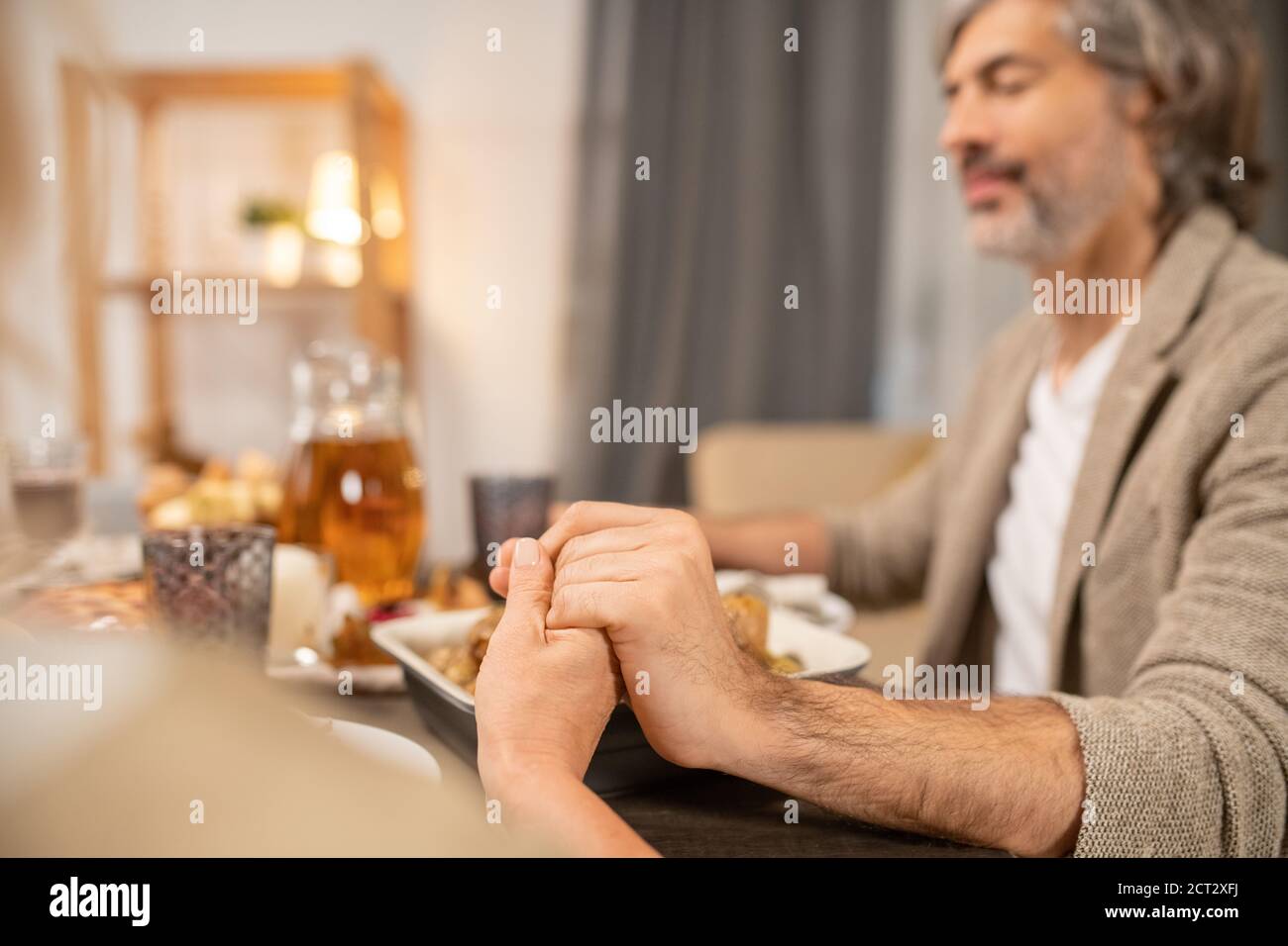Manos de un hombre maduro con los ojos cerrados y su esposa orando por mesa servida Foto de stock