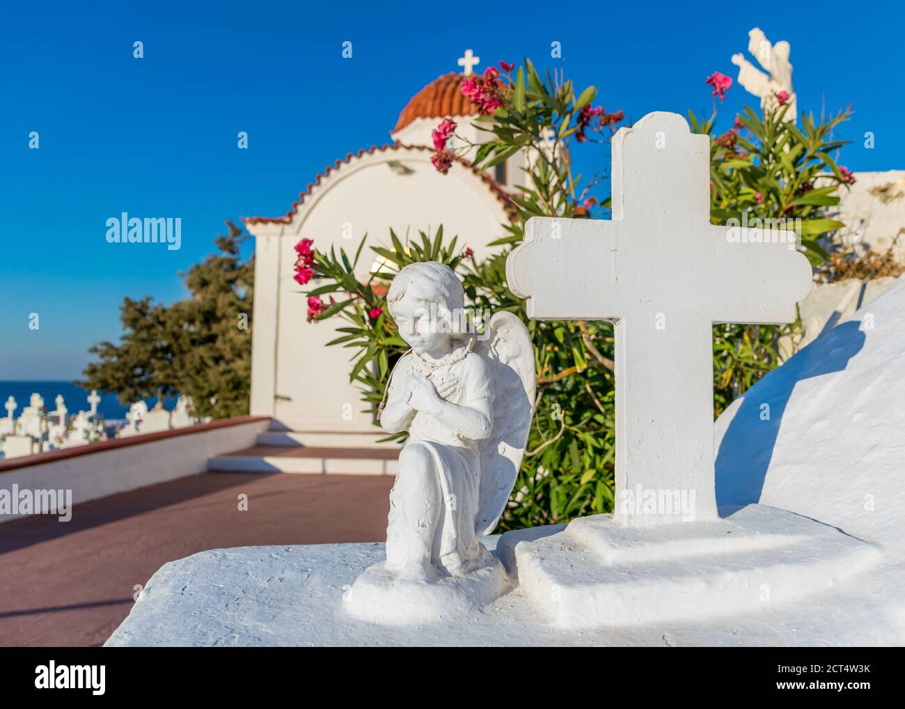 Estatua de ángel y cruz rezando con iglesia tradicional con cúpula roja en el fondo, isla Karpathos, Grecia Foto de stock