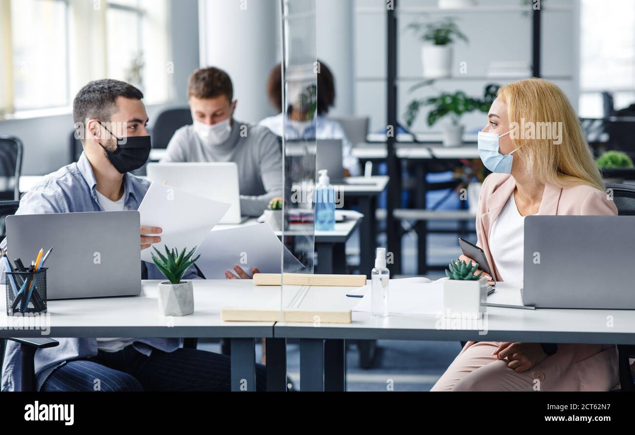 Nuevo normal y comunicarse con protección. El hombre milenario y la mujer en máscaras protectoras trabajan con documentos en vidrio protector Foto de stock