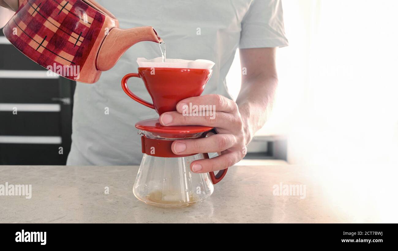 Café casero. Pourver. Un hombre hace una cerveza en casa v 60. Vierte agua  caliente en un gotero de cerámica roja con un filtro y el café gotea en un  servidor de