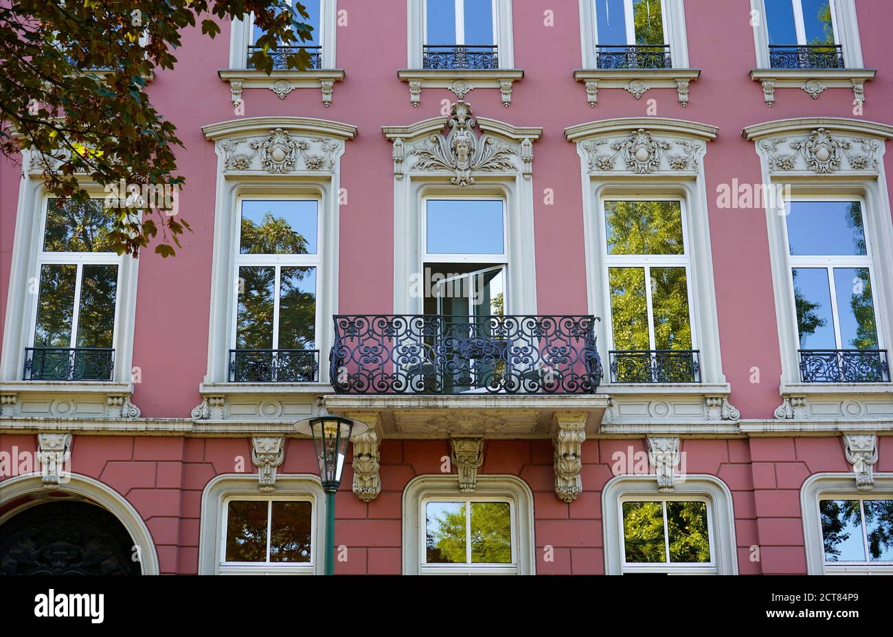 Romántica fachada de color rosa de la casa con balcón de metal y marcos de ventanas blancas. Cielo azul y árboles que se reflejan en los cristales de la ventana. Foto de stock