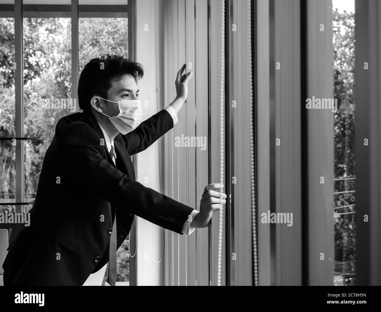 Hombre de negocios asiático solitario y cansado en traje con máscara médica de pie y mirando por la ventana de cristal en la oficina en blanco y negro estilo Fotografía stock -