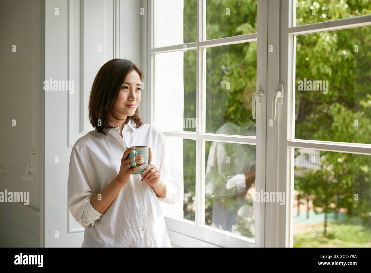 joven mujer asiática de pie junto a la ventana en casa sosteniendo un taza de café que se ve feliz y contento Foto de stock