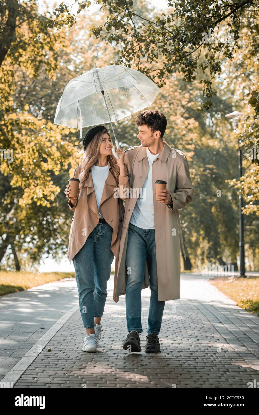 pareja abrigos de trinchera caminando bajo paraguas y sosteniendo papel tazas con café llevar de stock - Alamy