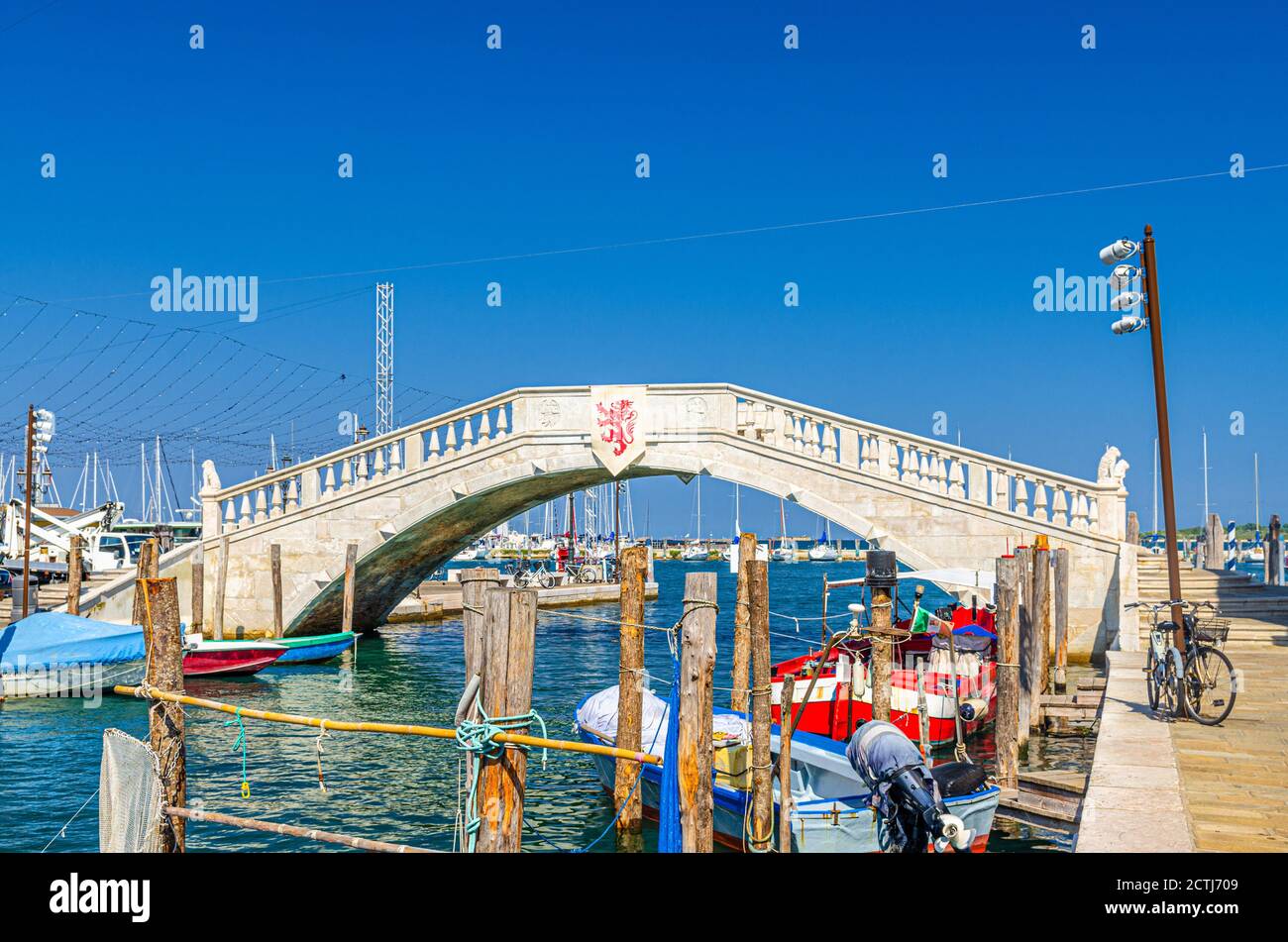 Puente de piedra Ponte di Vigo a través del canal de agua vena con coloridos barcos cerca del terraplén del muelle en el centro histórico de la ciudad de Chioggia, cielo azul de fondo en el día de verano, Región Veneto, Norte de Italia Foto de stock