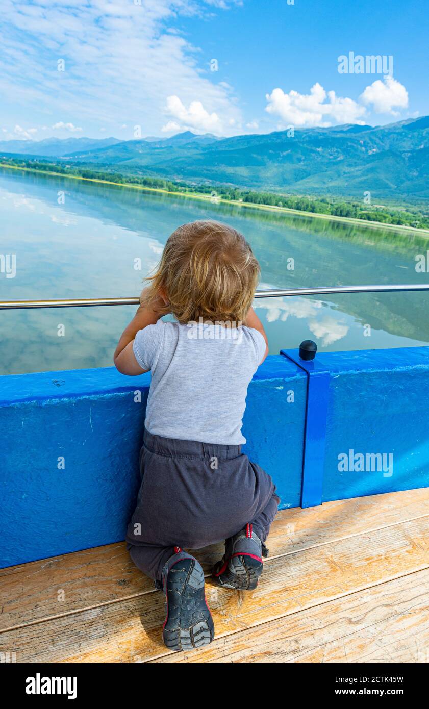 Niño arrodillado frente a la barandilla mirando la vista panorámica del lago Kerkini, Macedonia, Grecia Foto de stock