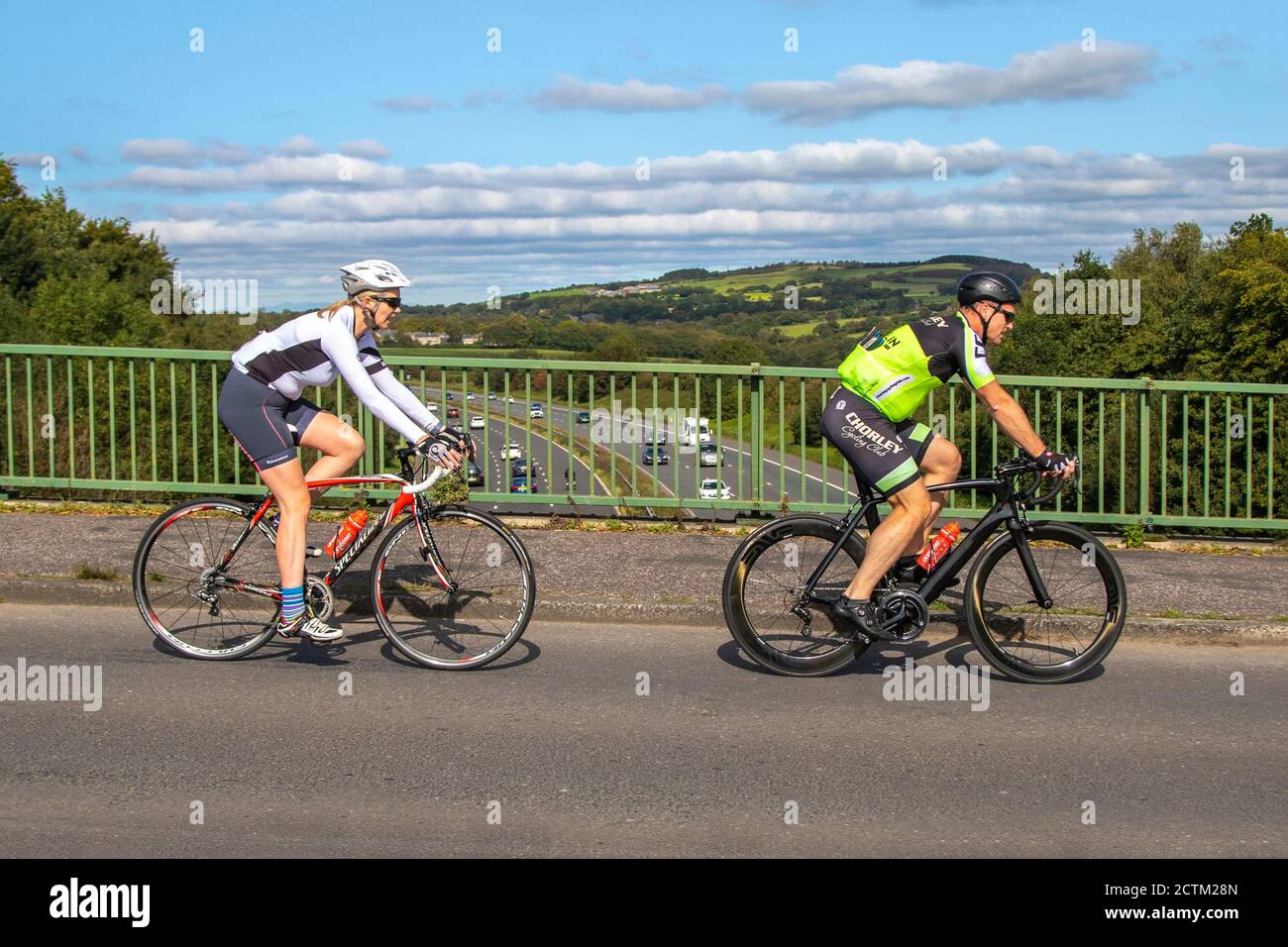 Hombre y mujer ciclistas montando bicicleta deportiva especializada cruzando por encima de la autopista M61 en un puente, cerca de Manchester Reino Unido Foto de stock